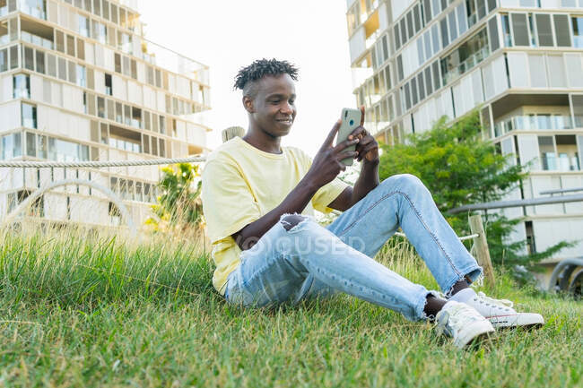 Повне тіло усміхненого молодого чорного чоловіка у світло-жовтій сорочці та блакитних джинсах та кросівках, що сидять на траві та переглядають смартфон — стокове фото