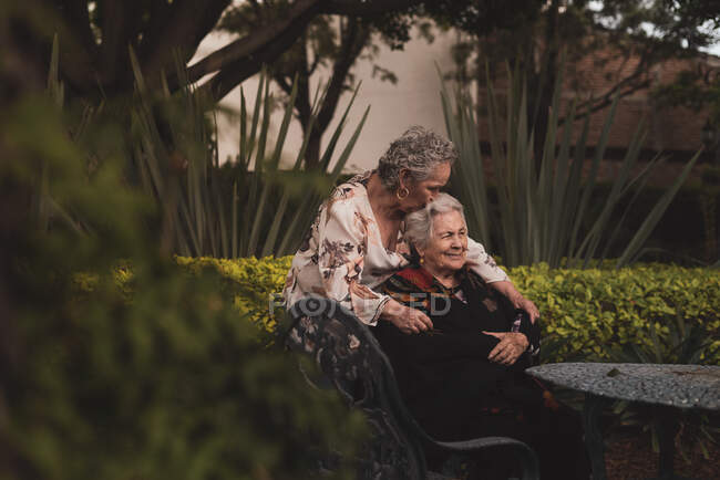 Feliz anciana abrazando y besando a la hermana en la cabeza y luego sonriendo mientras pasan tiempo en el jardín juntos - foto de stock