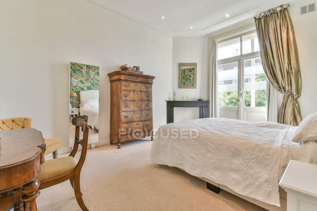 Interior de amplio dormitorio con cómoda cama suave y muebles de madera de estilo clásico y sillón en apartamento ligero - foto de stock