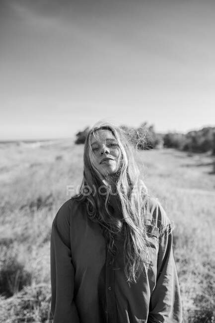 Black and white of anonymous female in shirt standing on grassy meadow under cloudless sky in summer looking at camera — Stock Photo