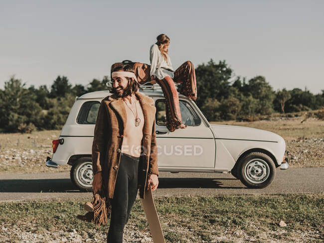 Pareja hippie alegre en trajes de estilo boho pasar tiempo juntos en el campo con el coche blanco viejo temporizador durante el viaje en la naturaleza - foto de stock