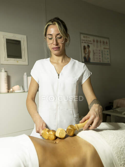 Massagista feminina com massageador de madeira fazendo massagem maderoterapia na barriga do cliente perto de toalhas no salão de spa leve — Fotografia de Stock