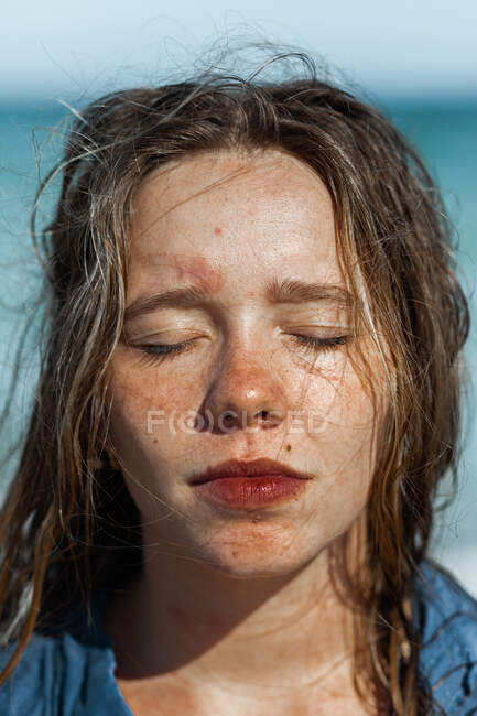 Female in wet shirt and with wet hair standing on beach near sea while enjoying summer day with eyes closed — Stock Photo