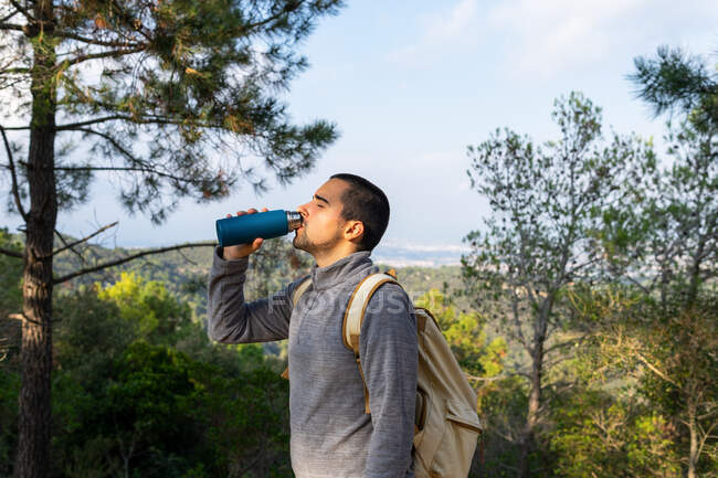 Jovem caminhante masculino hispânico barbudo em roupas casuais bebendo em exuberante floresta verde com garrafa térmica na mão no dia ensolarado — Fotografia de Stock