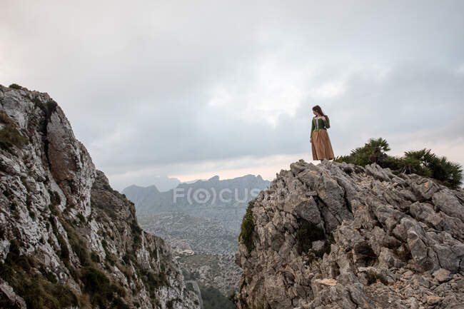 Вид збоку тіла мрійливої жінки в старомодному одязі, що стоїть на краю кам'яної скелі біля моря в туманній погоді — стокове фото