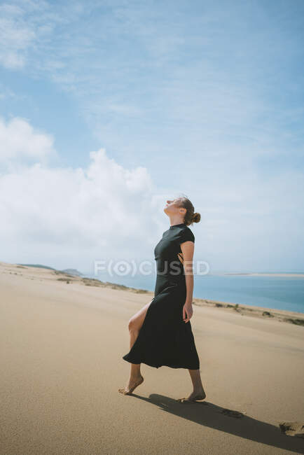 Full body side view of a female in dark dress looking at the sun with eyes closed on sandy dune in desert and in the background the sea — Stock Photo