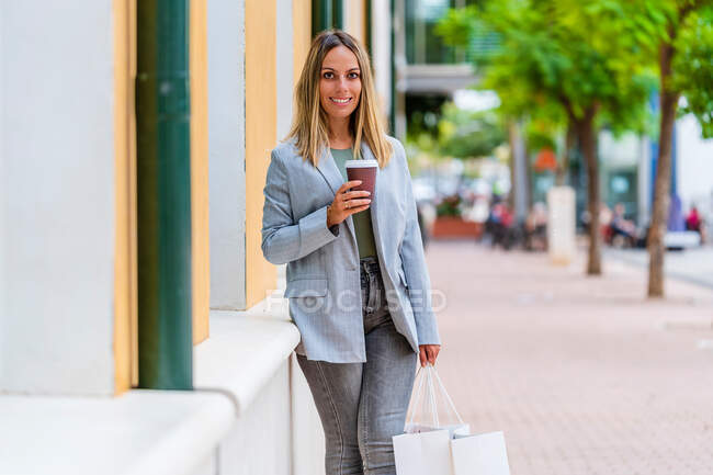 Фронтальный вид позитивной женщины в шикарном костюме с кофе, которая смотрит в камеру, держа в руках пару сумок для покупок — стоковое фото