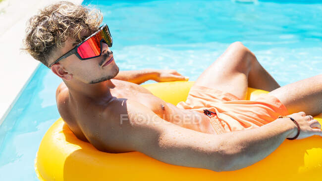 Homem calmo em calções de banho e óculos de sol banhos de sol no anel inflável amarelo na piscina com água clara no dia ensolarado de verão — Fotografia de Stock