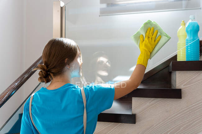 Back view of young professional cleaner in uniform and yellow rubber gloves wiping dust from glass of staircase while working in modern house in daylight — Stock Photo