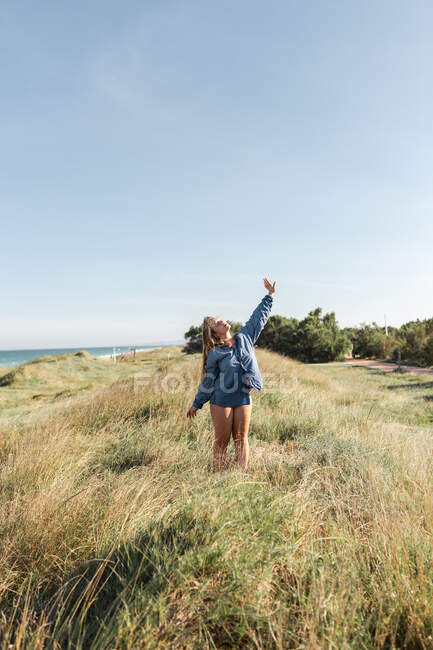 Blanco y negro de mujer anónima en camisa de pie con los brazos levantados en pradera cubierta de hierba bajo cielo despejado en verano con los ojos cerrados - foto de stock