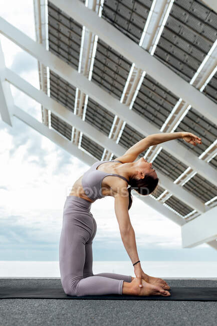 Ganzkörper-Seitenansicht einer entschlossenen Sportlerin, die sich darauf vorbereitet, während des Yoga-Trainings eine Ushtrasana-Haltung auf einer Matte in der Nähe von Solarzellen durchzuführen — Stockfoto