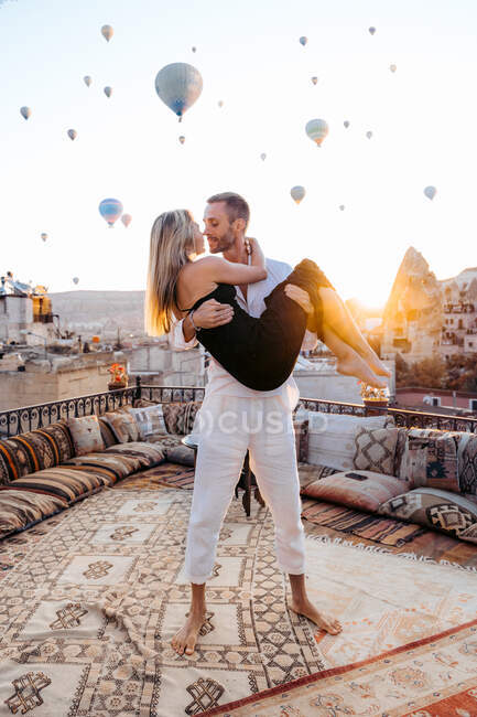 Full body of man holding girlfriend in hands while standing on terrace on background of hot air balloons in sunlight — Stock Photo