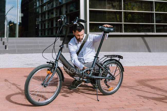 Полное тело взрослого бородатого мужчины в формальной одежде, закрепляющее заднюю цепь велосипедного колеса в парке в центральном районе — стоковое фото