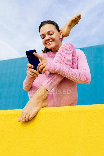 Позитивний стрункий жіночий обмін повідомленнями на мобільному телефоні під час сидіння у варіанті пози Seated Cradle, під час виконання вправ з йоги на чорному та жовтому фоні. — стокове фото