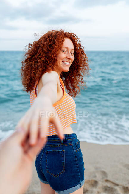 Вид на беззаботную молодую женщину-путешественницу с вьющимися рыжими волосами, держащую за руку бойфренда урожая на песчаном пляже, омытом синим морем — стоковое фото