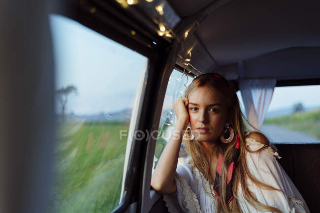 Confiada hermosa chica rubia apoyada en la ventana dentro de una furgoneta vintage mirando a la cámara - foto de stock