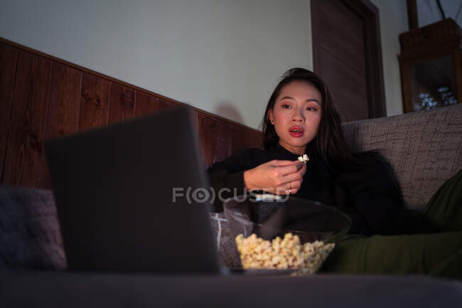 Молодая азиатка в повседневной одежде сидит на диване и ест попкорн дома, смотрит кино на ноутбуке — стоковое фото