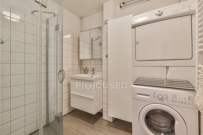 Стеклянная душевая кабина возле современной белой стиральной машины в просторной ванной комнате с керамической раковиной на стене с зеркалом в квартире — стоковое фото