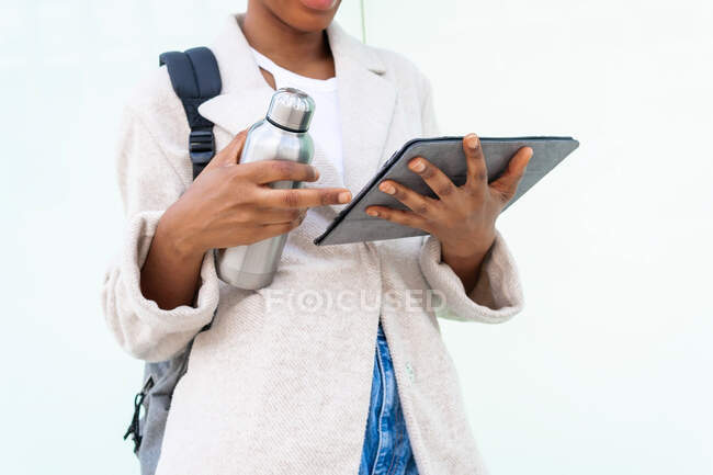 Невідома афроамериканська студентка з рюкзаком і термос гарячого напою серфінг інтернет на планшеті на білому тлі — стокове фото