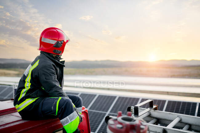 Anonymous pensive adult male wearing protective hardhat and protective suit while sitting on top of fire engine and looking forward at solar panels in rural farm with clear sky on background — Stock Photo