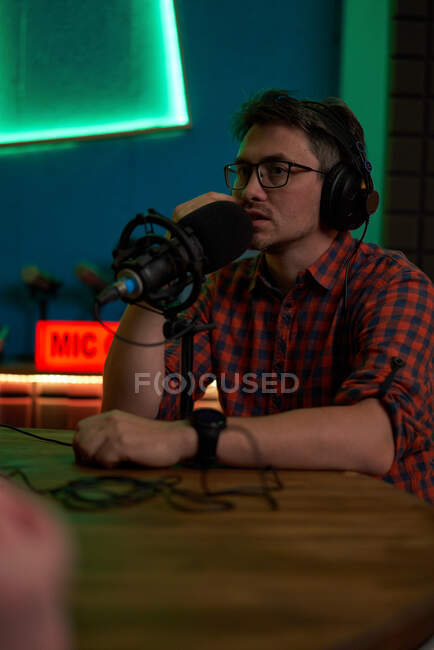 Joven presentador de radio masculino con camisa a cuadros y auriculares sentados en la mesa con micrófono y comunicándose con su colega anónimo durante la grabación de podcast en el estudio - foto de stock