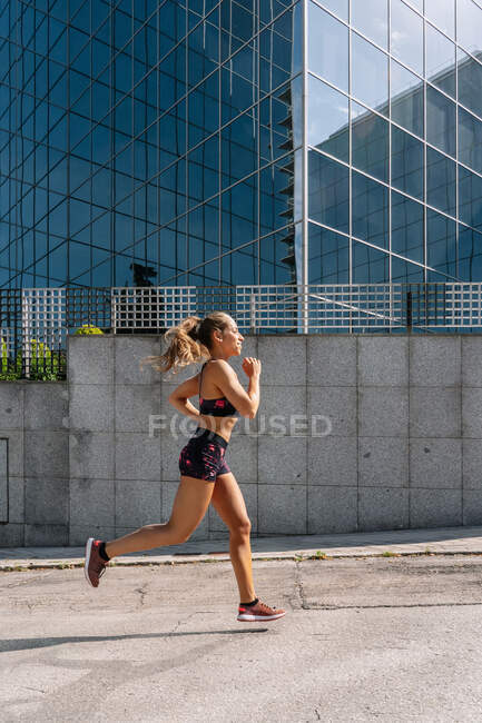 Повний вигляд стрункої активної жінки - бігуна в спортивному одягу біжить по покритій покришкою проїжджій дорозі в сучасному районі міста. — стокове фото