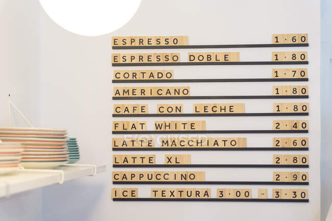 Доска меню с различными кофейными напитками и ценами на белой стене возле прилавка с посудой в современном светлом кафе с светящимся светом — стоковое фото
