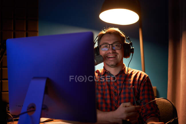 Side view of focused young male in checkered shirt and eyeglasses using computer and speaking in mic while recording podcast in dark studio — Stock Photo