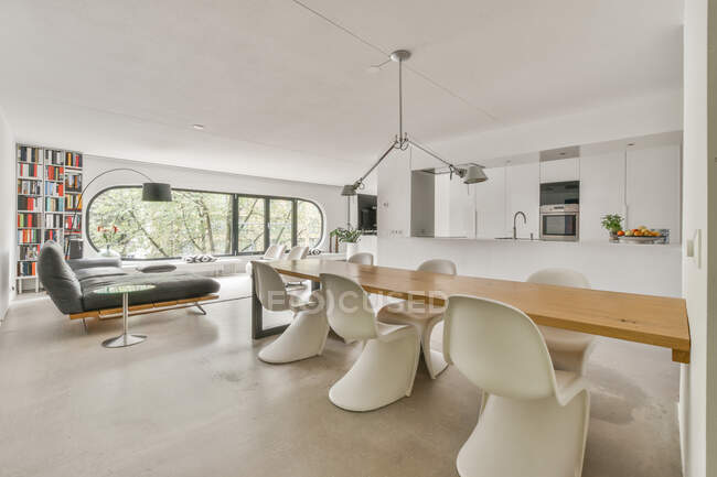 Quarto contemporâneo e cozinha interior com sofá e mesa sob lâmpadas contra janela e estante em casa — Fotografia de Stock