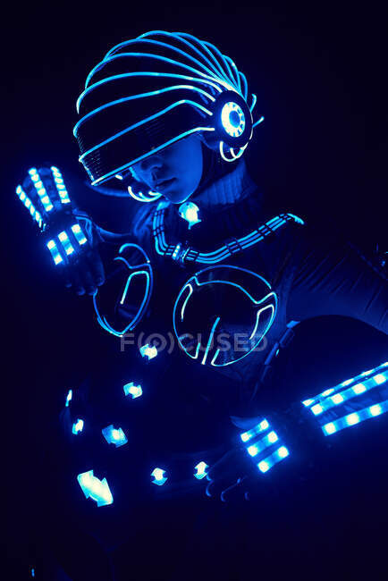 Persona sin rostro en traje brillante contemporáneo de cyborg espacio con iluminación de neón y casco de pie sobre fondo negro en estudio oscuro - foto de stock