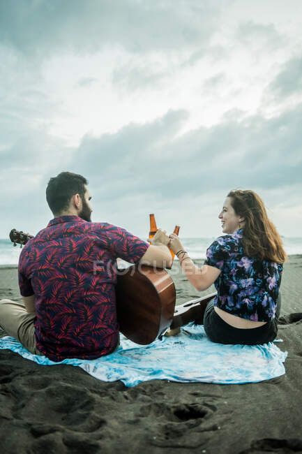 Vista trasera de un par de músicos positivos sentados con guitarras y botellas de cerveza mientras están sentados en la playa de arena cerca del océano durante el día - foto de stock