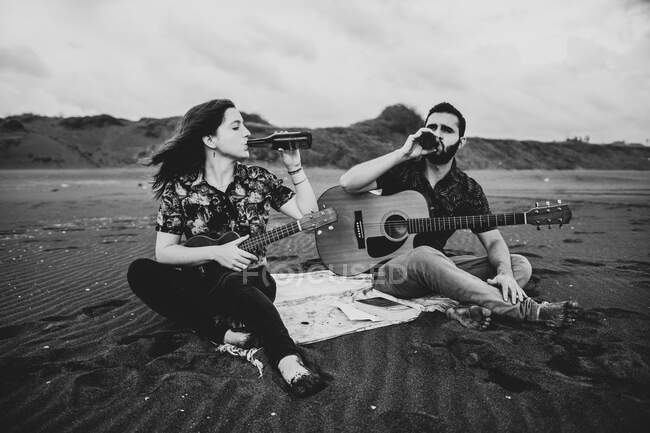 Noir et blanc de femme calme avec ukulélé assis et boire de la bière avec un ami guitariste sur la rive sablonneuse dans la nature en plein jour — Photo de stock