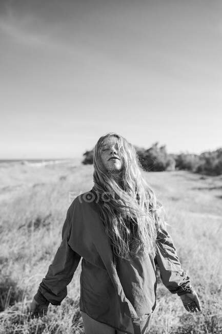 Black and white of anonymous female in shirt standing on grassy meadow under cloudless sky in summer with eyes closed — Stock Photo