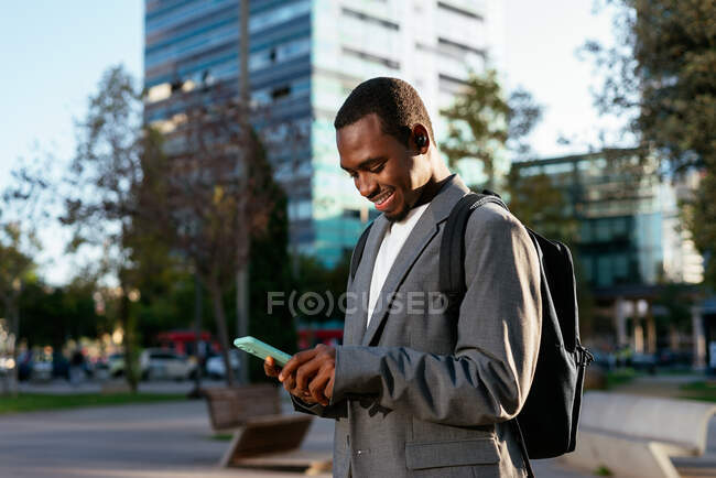 Homme d'affaires afro-américain souriant en tenue formelle avec sac à dos naviguant sur le téléphone mobile dans le centre-ville et regardant vers le bas avec sourire dents — Photo de stock