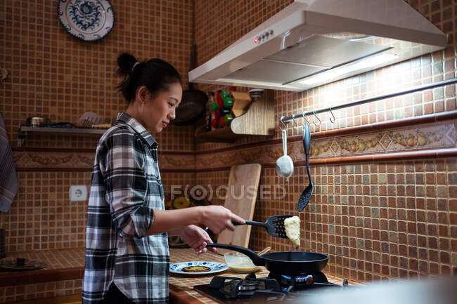 Бічний погляд на сфокусовану молоду азіатку в повсякденному одязі куховарські млинці з використанням спатули, стоячи на кухні. — стокове фото