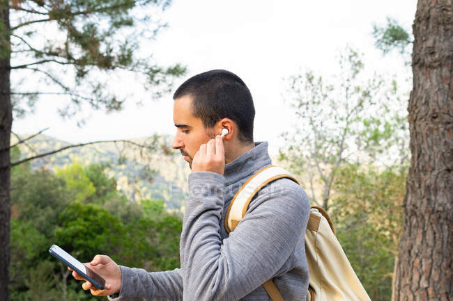 Бічний погляд на серйозного молодого іспаномовного чоловіка в повсякденному одязі і рюкзак, який слухає музику через бездротові навушники, використовуючи смартфон під час подорожі в лісі. — стокове фото
