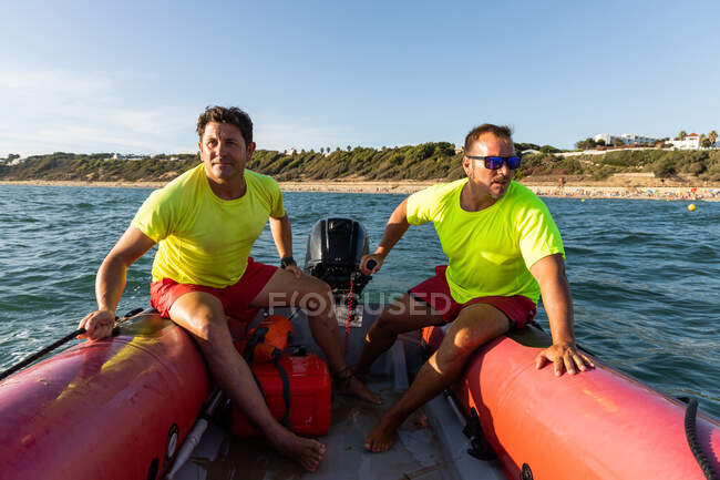 Full length of barefoot lifeguards sitting on inflatable motorboat floating on rippling blue sea and controlling safety on sea — Stock Photo