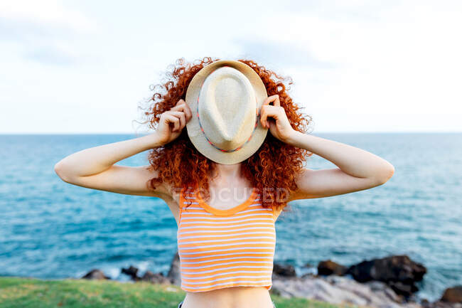 Mulher irreconhecível com longos cachos de gengibre cobrindo rosto com chapéu na costa do mar azul — Fotografia de Stock