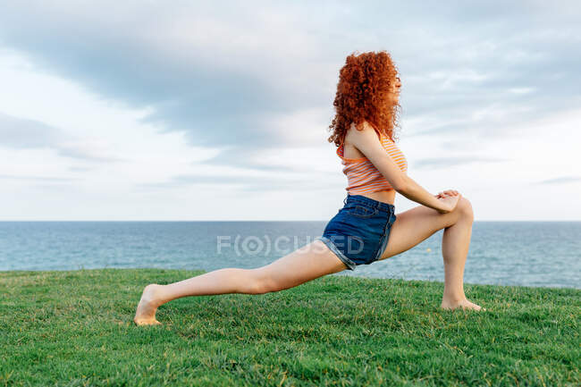 Full body side view of positive curly haired female practicing high lunge yoga posture on grassy lawn on seashore — Stock Photo