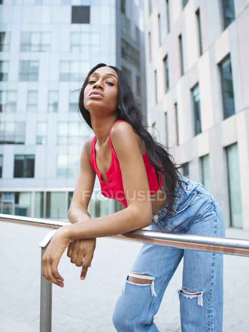 Mulher afro-americana confiante em roupas elegantes olhando para a câmera enquanto se inclina sobre trilhos na cidade com modernos edifícios de vários andares — Fotografia de Stock