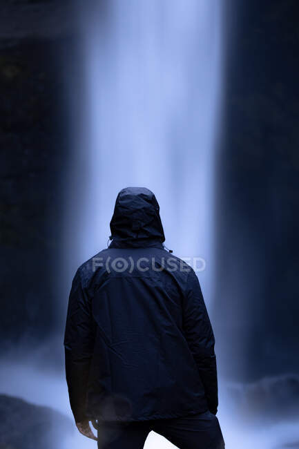 Back view of anonymous male traveler in warm outfit standing and admiring picturesque rapid waterfall — Stock Photo