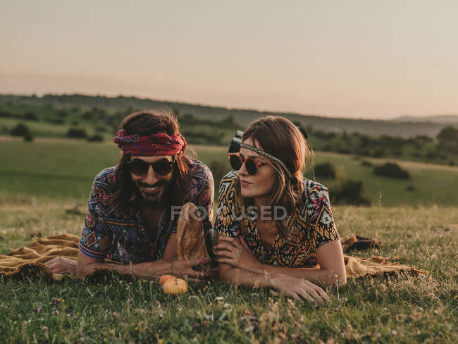 Casal Hippie com óculos de sol olhando para longe enquanto deitado no prado gramado na natureza ao pôr do sol tempo — Fotografia de Stock