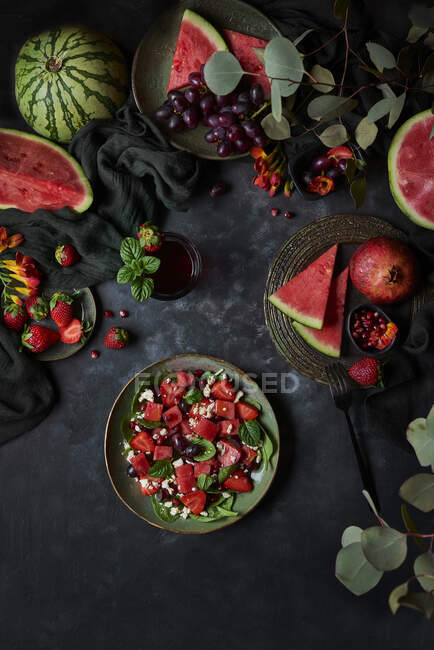 Вид зверху тарілка смачного салату з червоним кавуном і полуницею, розміщена на чорному тлі зі стиглим гранатом і виноградом — стокове фото