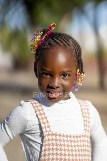 Felice ragazza afroamericana con acconciatura in abito elegante in piedi e guardando la fotocamera contro sfondo sfocato sulla strada in giornata di sole — Foto stock