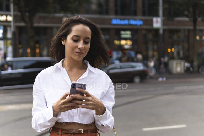 Confident female in stylish outfit using smartphone while standing on city street near road with cars — Stock Photo