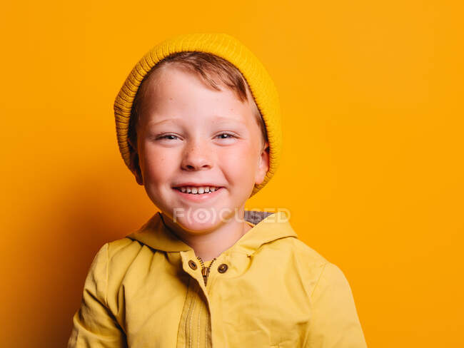 Niño feliz en chaqueta de impermeable amarillo vivo y gorro sombrero riendo y mirando a la cámara contra el fondo amarillo en el estudio - foto de stock