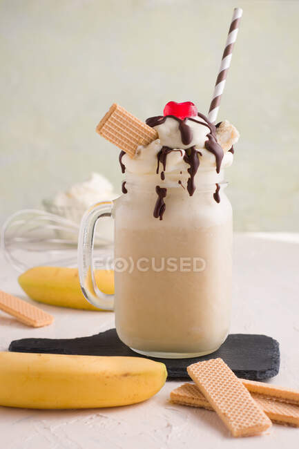 Glass jar of sweet banana split milkshake topped with whipped cream waffles chocolate and cherry on cutting board — Stock Photo