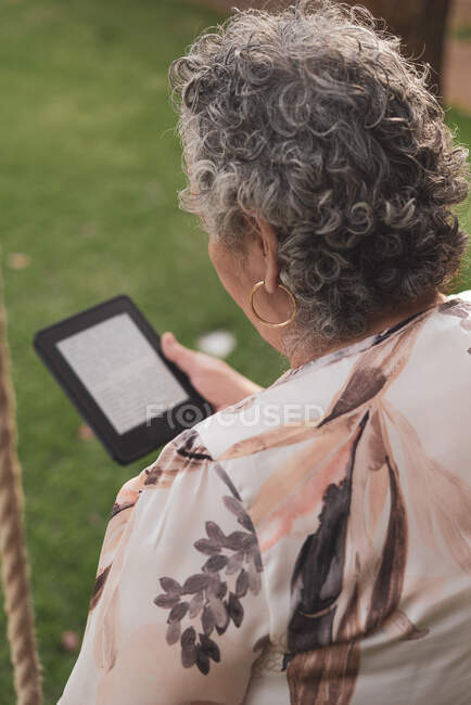 Vista posteriore della signora anziana che indossa la camicetta seduta nel parco e legge il libro elettronico — Foto stock