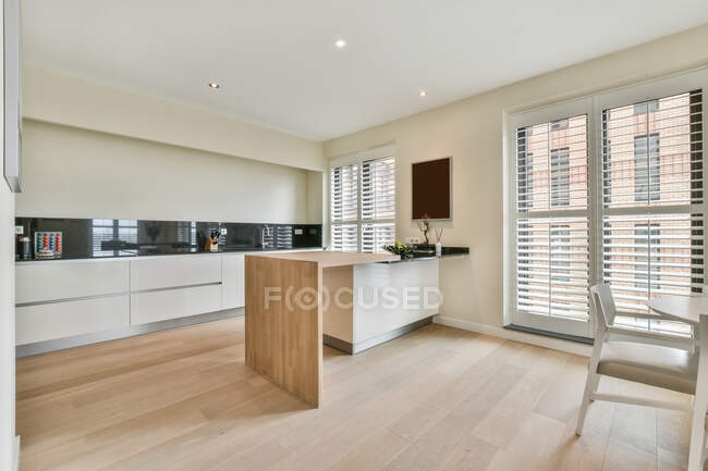 Bancone in legno posizionato vicino a armadi bianchi con forniture in spaziosa zona cucina in elegante appartamento luminoso con finestre e sedia — Foto stock
