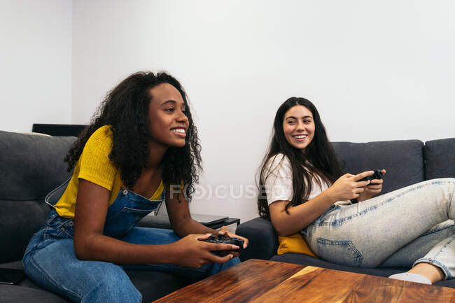 Позитивні багаторасові друзі-жінки сидять на дивані і грають у відеоігри, проводячи час разом вдома — стокове фото